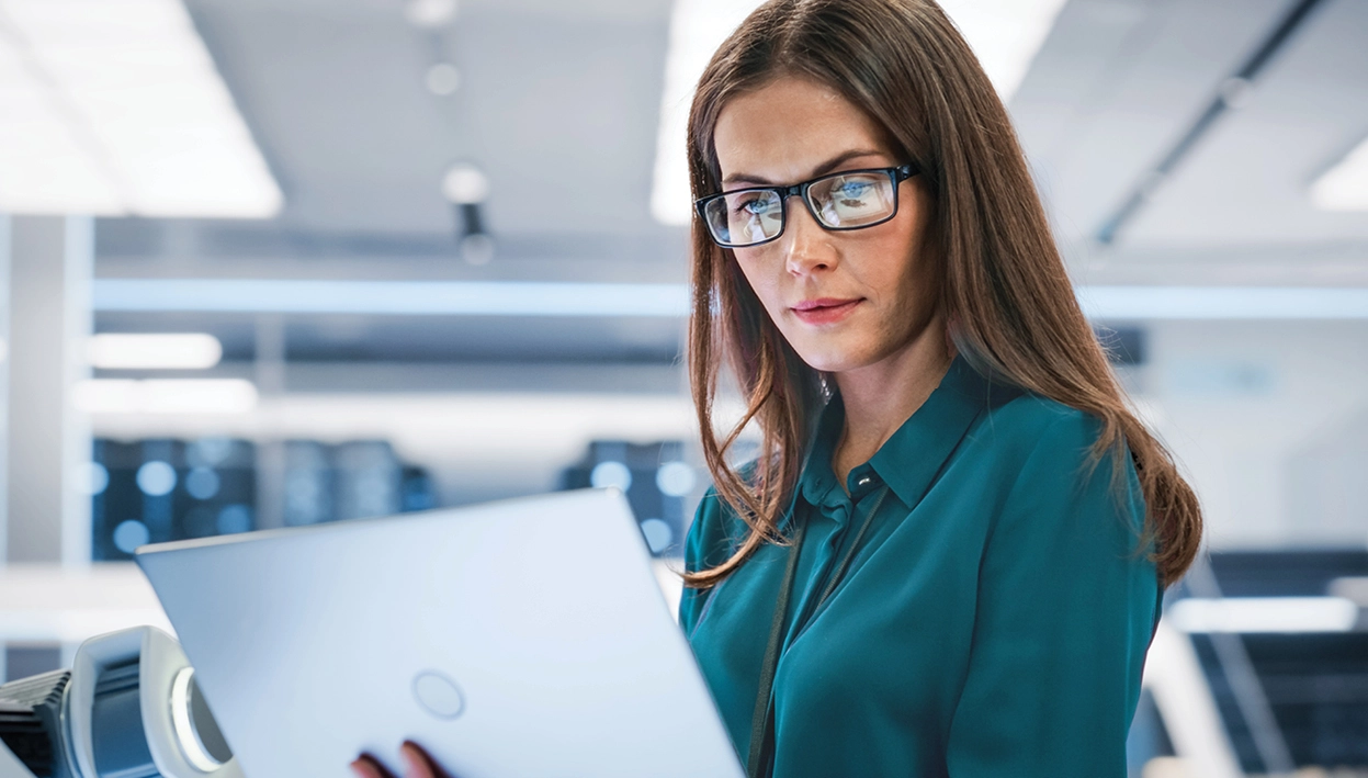 A woman wearing glasses is focused on a laptop, engaged in IT support tasks.
